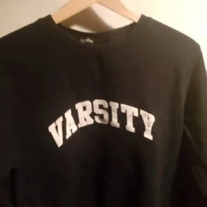 Svart sweatshirt med texten 'Varsity' - Säljer en svart sweatshirt från Brother med texten 'Varsity' i vitt på framsidan. Perfekt till både jeans och joggers. Tröjan har långa ärmar och en klassisk rund halsringning.