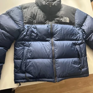 Blå och svart dunjacka från The North Face - Säljer en snygg blå och svart dunjacka från The North Face. Jackan har en dragkedja framtill och är perfekt för kyliga dagar. Den är långärmad och har en klassisk pufferdesign med logotyp både i fram och bak💕🙌🏼