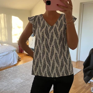 Blåvit mönstrad blus - Snygg blus med blåvitt mönster och v-ringning. Blusen har korta ärmar med en lätt volangdetalj. Perfekt för en stilren look.
