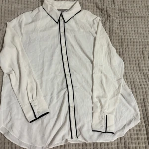 Vit blus med svarta detaljer - Elegant vit blus med svarta kantdetaljer längs krage och knäppning. Långärmad design med knappar framtill och vid ärmslut. Perfekt för en stilren look. Om ni vill se den på, skriv gärna!