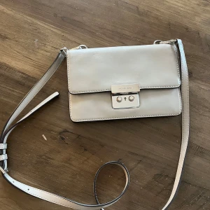 Beige axelväska från Michael Kors - Snygg beige axelväska från Michael Kors med justerbar axelrem och stilren design. Väskan har ett metallspänne framtill och flera fack inuti för smidig organisering. Finns tecken på användning såsom repor på metallspännet. 