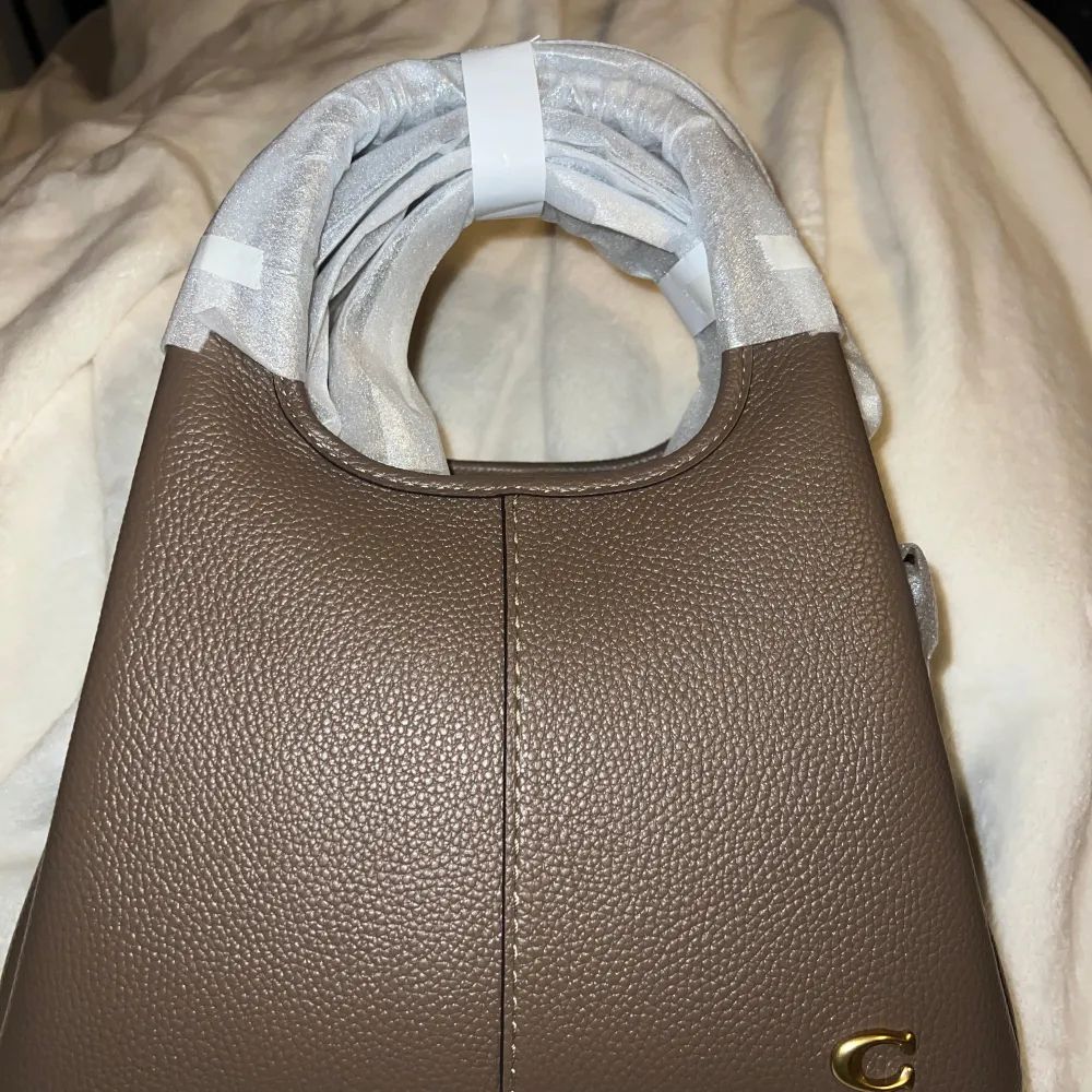 Snygg brun handväska från Coach i skinn med en stilren design. Väskan har en guldlogga på framsidan och bekväma handtag. Perfekt för att ge din outfit en elegant touch.. Laukut & Käsilaukut.