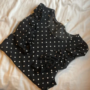 Svart prickig blus - Snygg svart blus med vita prickar och långa ärmar. Blusen har en rund halsringning och volangdetaljer på ärmarna som ger en extra touch. Perfekt för att piffa upp vardagslooken!