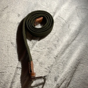 Grönt flätat bälte med bruna läderdetaljer - Snyggt grönt flätat bälte med bruna läderdetaljer och silverfärgat spänne. Perfekt för att ge en stilren touch till din outfit. Bältet har en unik flätad design som ger det en modern känsla.