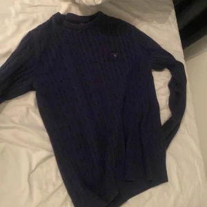 Mörkblå stickad tröja från GANT - Säljer en snygg mörkblå stickad tröja från GANT med kabelstickat mönster. Tröjan har en rund halsringning och långa ärmar. Perfekt för kyligare dagar!