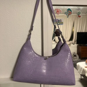 Lila axelväska med krokodilmönster - Snygg lila axelväska med krokodilmönster och dragkedja. Perfekt för att ge en färgklick till din outfit. Väskan har en stilren design och är lätt att bära med sig.