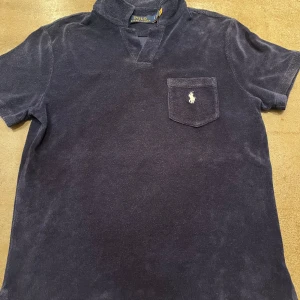 Mörkblå frotté t-shirt från Polo Ralph Lauren - Säljer en mörkblå frotté t-shirt från Polo Ralph Lauren med korta ärmar och en bröstficka med broderad logga. Perfekt för en avslappnad stil.