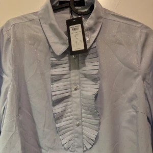 Ljusblå skjorta från Vero Moda - Elegant ljusblå skjorta från Vero Moda med subtila prickar och volangdetaljer framtill. Skjortan har långa ärmar och klassisk krage, perfekt för en stilren look.