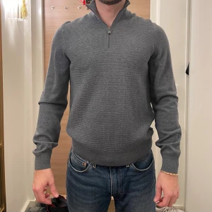 Grå tröja med dragkedja från BOSS - Snygg grå tröja från BOSS med halv dragkedja och ribbade muddar. Perfekt för en stilren look. Tröjan har en klassisk passform och är tillverkad i ett mjukt material. Originalpris: 1999 kr