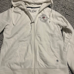 Vit huvtröja från Polo Club - Säljer en vit huvtröja från Polo Club Saint Vincent med dragkedja och kängurufickor. Tröjan har en broderad logga på bröstet och är tillverkad i mjukt material. Perfekt för en avslappnad stil.
