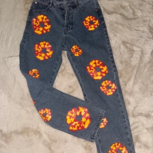 Offret Denim Tears - Unika blå jeans från Levi's med färgglada blommönster i rött och gult. Byxorna har en klassisk femficksdesign och knappgylf. Perfekta för att ge din outfit en färgglad touch.