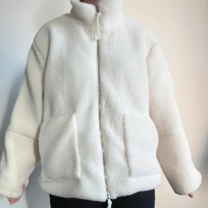 Vit teddyjacka med dragkedja - Mysig vit teddyjacka med dragkedja och stora fickor framtill. Perfekt för kyliga dagar med sin fluffiga och varma design. Jackan har en hög krage och en avslappnad passform.