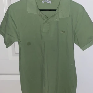 Grön pikétröja från Lacoste - Snygg grön pikétröja från Lacoste med klassisk krage och korta ärmar. Perfekt för en avslappnad stil. Den har en liten broderad logotyp på bröstet. Tröjan är på ett bra skick helt oanvänd så den fläcken på chockar mig också med den syns bara om du blir filmad med flash annars så syns den inte. 