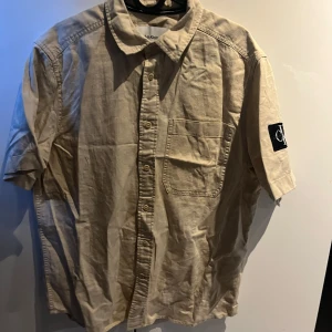 Beige kortärmad skjorta från Calvin Klein - Snygg beige skjorta från Calvin Klein med korta ärmar och klassisk krage. Skjortan har en bröstficka och knappar framtill. Perfekt för en avslappnad stil. 