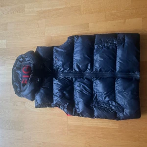 Blå dunväst med huva - Snygg blå dunväst med huva och dragkedja. Västen har en cool design med röd och vit detalj på baksidan. Perfekt för kyligare dagar.