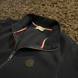 Marinblå tröja med dragkedja från Moncler - Snygg marinblå tröja från Moncler med dragkedja och långärmad design. Tröjan har en diskret logga på bröstet och röd-vit detalj vid kragen. Perfekt för en stilren look.