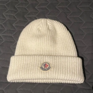 Moncler mössa - Snygg vit ribbad mössa från Moncler med broderad logotyp framtill. Perfekt för att hålla värmen med stil under kyliga dagar.
