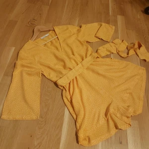 Jumpsuit- Byxdress i shortsmodell från Na-kd - Charmig gul klänning med vita prickar från Na-kd. Klänningen har en v-ringad design och trekvartsärmar. Den är försedd med ett knytbälte i midjan för en smickrande passform.