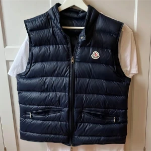 Svart dunväst från Moncler - Snygg svart dunväst från Moncler med dragkedja och två praktiska fickor framtill. Perfekt för kyligare dagar och ger en stilren look. Västen har en diskret logga på bröstet.