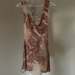 Mönstrad klänning med öppen rygg - Säljer en kort klänning med ett vackert paisleymönster i rosa/brunt. Klänningen har en öppen rygg med knytband och är ärmlös. Köpt på ASOS. Aldrig använd