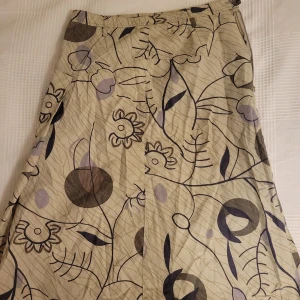 Mönstrad kjol från Green House - Säljer en beige blommig kjol från Green House i storlek 36. Kjolen är tillverkad av 100% bomull och har ett vackert blommönster i lila och brunt. Den har en dragkedja i sidan för enkel på- och avtagning.