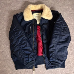 Blå bomberjacka från Alpha Industries - Säljer en snygg blå bomberjacka från Alpha Industries med en mysig pälsliknande krage. Jackan har en quiltad röd insida och dragkedja framtill. Perfekt för kyligare dagar! Size M men passar också S, skick 10/10