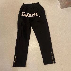 Svarta mjukisbyxor med dragkedja och tryck fram - Svarta mjukisbyxor från Dozani med elastisk midja och dragkedjor vid bensluten. Perfekta för en avslappnad stil.