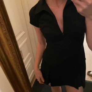 Svart kortklänning med dragkedja - Snygg svart kortklänning med dragkedja framtill och korta ärmar. Perfekt för en stilren look. Klänningen har en figurnära passform som framhäver silhuetten.