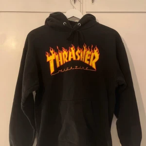 Svart hoodie från Thrasher - Säljer en svart hoodie från Thrasher med den ikoniska loggan i eldflammor på bröstet. Perfekt för skate-entusiaster eller streetwear-fans. Hoodien har en klassisk passform med känguruficka och justerbar huva.