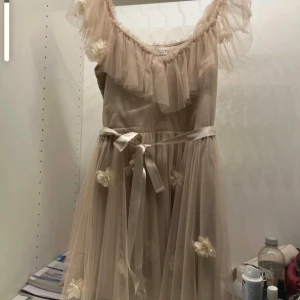 Ida Sjöstedt Champagnefärgad klänning  - Elegant champagnefärgad klänning med volangdetaljer och blommor. Klänningen har en offshoulder-design och ett knytband i midjan. Perfekt för speciella tillfällen. Använd en gång 