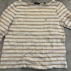 Randig tröja från Gant - Säljer en stilren randig tröja från Gant i beige och vit. Tröjan har ett litet broderat emblem på bröstet. Perfekt för en avslappnad look. Den är storlek M men är mer som s
