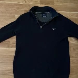 Mörkblå tröja med dragkedja från Gant - Säljer en stilren mörkblå tröja från Gant med dragkedja vid halsen. Tröjan har långa ärmar och en liten Gant-logga på bröstet. Perfekt för en avslappnad men snygg look.