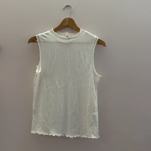 Vit ärmlös topp - Säljer en stilren vit ärmlös topp med en ribbad textur. Perfekt för varma dagar eller som ett lager under en jacka. Toppen har en enkel design med en rund halsringning och lätt vågiga kanter. är det storlek L men sitter väldigt tajt så skulle passa S eller M