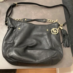 Svart axelväska från Michael Kors - Snygg svart axelväska i skinn från Michael Kors med guldfärgade detaljer och en fransig tofs. Väskan har en kedja och dragkedja för säker förslutning. Perfekt för att ge din outfit en lyxig touch.