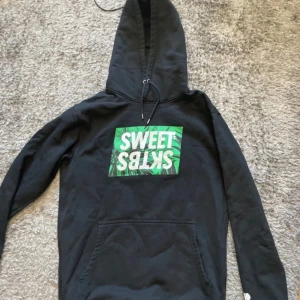 Svart hoodie från Sweet SKTBS - Säljer en svart hoodie från Sweet SKTBS med en grön och vit logga på framsidan. Den har en klassisk känguruficka och justerbar huva med dragsko. Perfekt för en avslappnad stil.