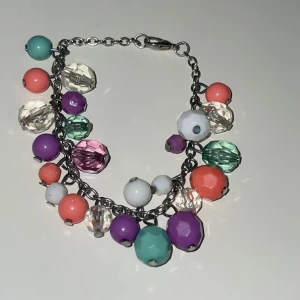 Färgglatt armband med pärlor - Charmigt armband med stora färgglada pärlor i turkos, lila, rosa, vit och grön. Pärlorna är fästa på en silverfärgad kedja med ett enkelt lås. Perfekt för att ge en färgklick till din outfit! Helt oanvänt!!