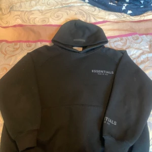 Svart hoodie från Essentials - Säljer en svart hoodie från Essentials med texten 'Fear of God' tryckt på bröstet och ärmen. Den har en klassisk design med huva och en stor ficka framtill. Perfekt för en avslappnad stil.