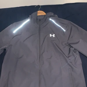 Svart jacka från Under Armour - Säljer en svart jacka från Under Armour med reflekterande detaljer på axlarna och dragkedja framtill. Jackan har en sportig look med logotyp på bröstet och text längs sidan. Perfekt för träning eller vardagsbruk.