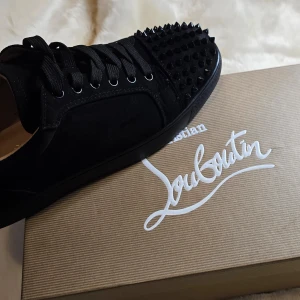 Svarta sneakers med nitar från Christian Louboutin - Snygga svarta sneakers från Christian Louboutin med nitar på tån och den ikoniska röda sulan. Skorna har snörning och är tillverkade i skinn, vilket ger en lyxig känsla. Perfekta för att ge din outfit en edgy touch. (Notera att dom är små i storleken så är du st 42 så passar dom) 