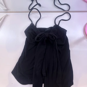 Svart axelbandslös topp från Gina Tricot - Säljer en stilren svart axelbandslös topp från Gina Tricot. Toppen har en söt rosett framtill och tunna axelband. Perfekt för en kväll ute eller en avslappnad dag. 