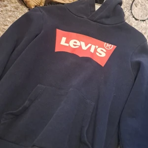 Mörkblå hoodie från Levi's - Säljer en snygg mörkblå hoodie från Levi's med det klassiska röda och vita logotyptrycket på bröstet. Tröjan har en praktisk magficka och en bekväm huva. Perfekt för en avslappnad stil.