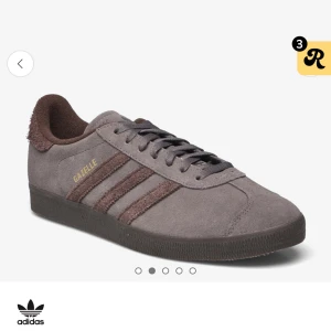 Grå/Bruna Adidas Gazelle sneakers. storlek:  44 1/3 - 1359 kr original pris.                                          Snygga grå Adidas Gazelle sneakers i mocka med de klassiska tre ränderna i mörkare ton. Skorna har snörning och en diskret guldfärgad Gazelle-logga på sidan. Perfekta för en stilren look.