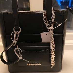 Svart axelväska från Steve Madden - Snygg svart axelväska från Steve Madden i syntetmaterial. Väskan har en stilren design med silverfärgade detaljer och en matchande plånbok. Perfekt för att hålla ordning på dina saker med stil. HELT NY! 