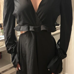 Svart omlottklänning - Snygg svart omlottklänning med volangdetaljer i midjan. Klänningen har långa ärmar och en v-ringad design som ger en elegant look. Perfekt för en stilren outfit.