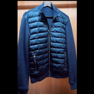 Stillren Cardigan  - Snygg blå jacka med quiltad framsida och stickade ärmar. Jackan har dragkedja framtill och två praktiska fickor med dragkedjor. Perfekt för kyligare dagar. Tveka inte på ställa frågor 