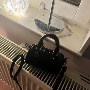 Svart handväska med gulddetaljer - Snygg svart handväska med gulddetaljer och dragkedja. Den har en stilren design med dubbla handtag och en avtagbar axelrem. Perfekt för att bära dina nödvändigheter med stil.