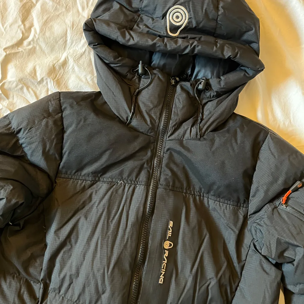 Snygg svart dunjacka från Sail Racing med huva och dragkedja. Perfekt för kalla dagar med sin vadderade design. Jackan har långa ärmar och en stilren look.. Takit.