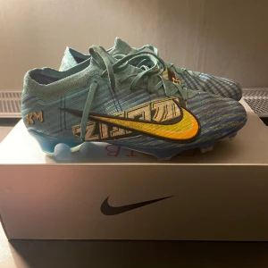 Nike Mercurial fotbollsskor - Snygga Nike Mercurial fotbollsskor i blått med gula och svarta detaljer. Skorna har snörning och en modern design med textur för bättre bollkontroll. Perfekta för fotbollsplanen.