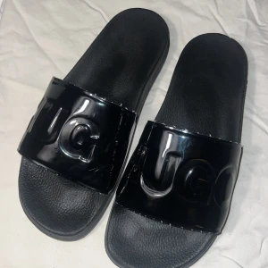 Svarta sandaler från Hugo BOSS - Snygga svarta sandaler från Hugo boss med en bred rem över foten. Remmen har en glansig finish med märkets logotyp i relief. Perfekta för en avslappnad stil.