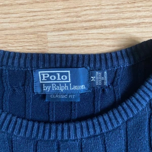  Polo Ralph Lauren - Säljer en stilren mörkblå kabelstickad tröja från Polo Ralph Lauren. Tröjan har en klassisk passform med rund hals och långa ärmar. Den ikoniska röda logotypen är broderad på bröstet. Perfekt för en elegant och avslappnad look.
